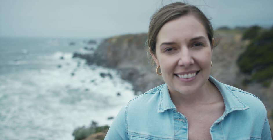 Woman stands near promontory overlooking ocean smiling