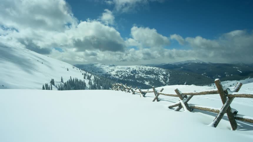 A snowy landscape with a fence with a mountain range the sky and clouds in the background