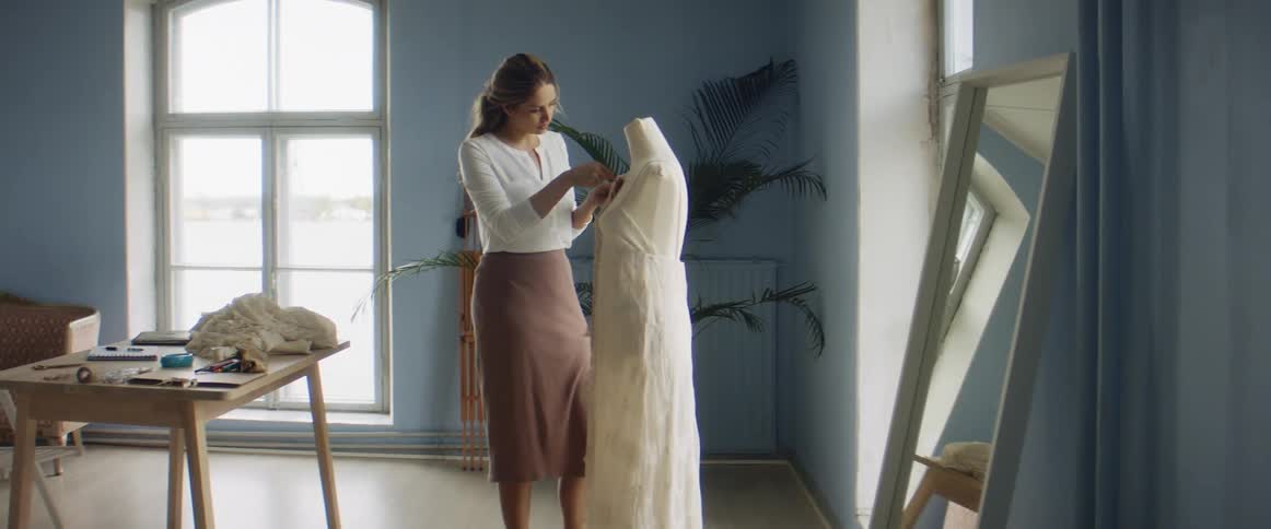 A woman stitches together a garment on a mannequin in front of a mirror indoors during the day