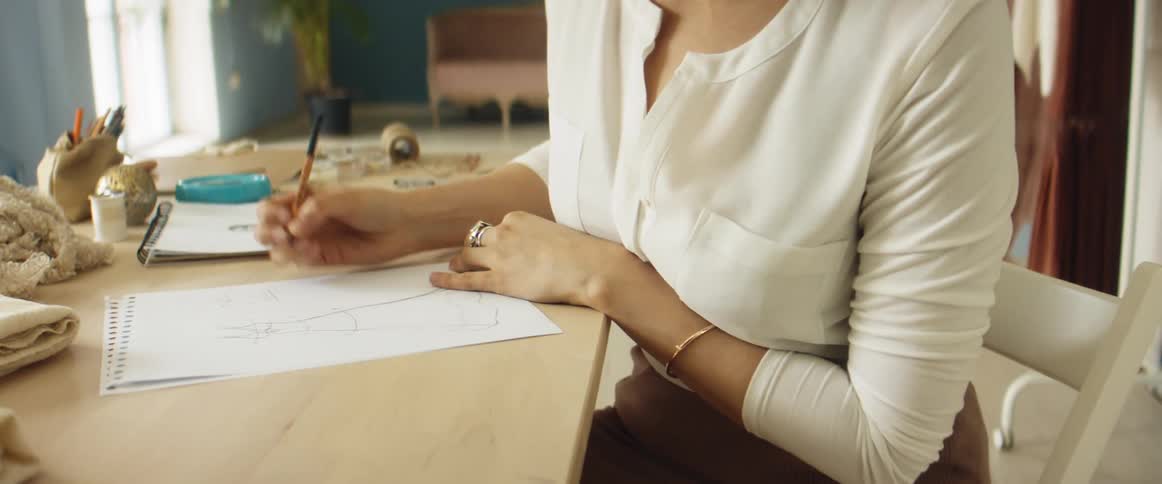 A woman in a room sitting on a chair at a desk drawing on a piece of paper with a pencil