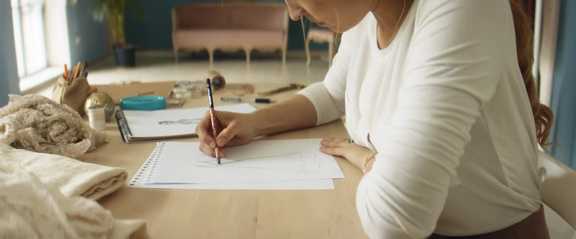 A woman is sitting on a desk and doing a pencil sketch