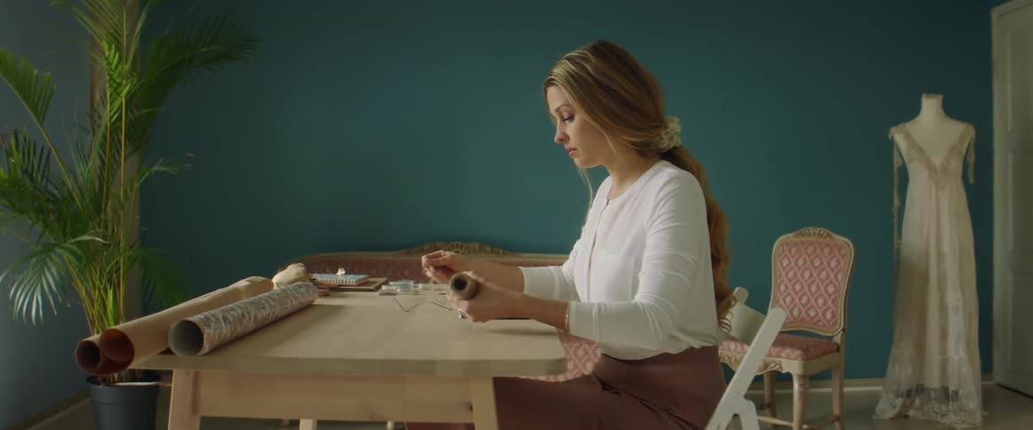 Designer sits at wooden table binding package with dress on dress form in background