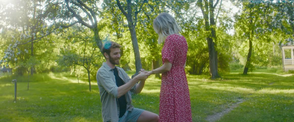 A man getting on one knee and proposing to a woman with trees the sky and clouds in the background