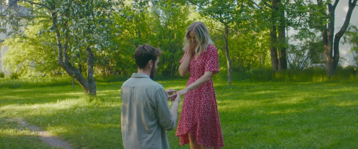 A man kneels to propose to a young woman and place a ring on her finger before standing and sharing a kiss with her