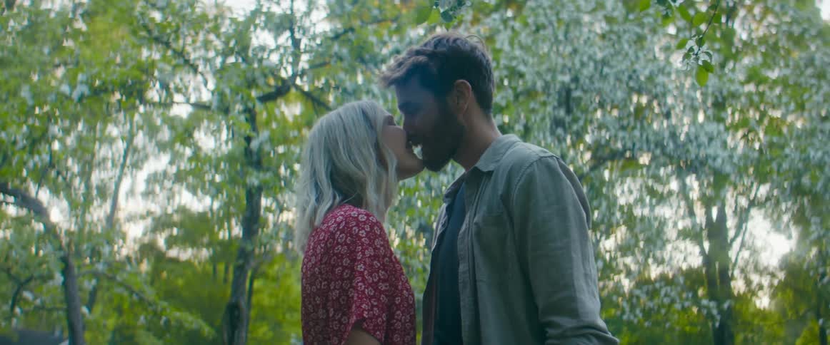 A woman moves closer to her boyfriend and kisses him as they stand in a forest