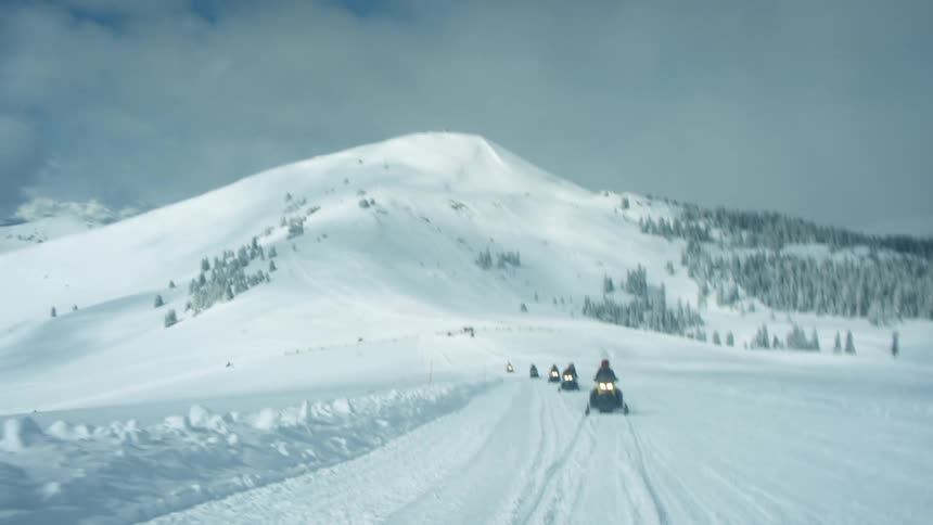 People on snowmobiles driving on a snowy landscape with a mountain range the sky and clouds in the background