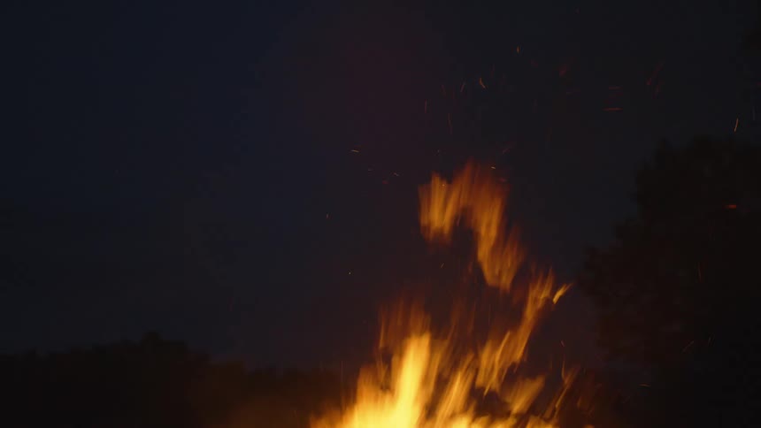 Fast moving and tall flames from a campfire kick embers into the night sky