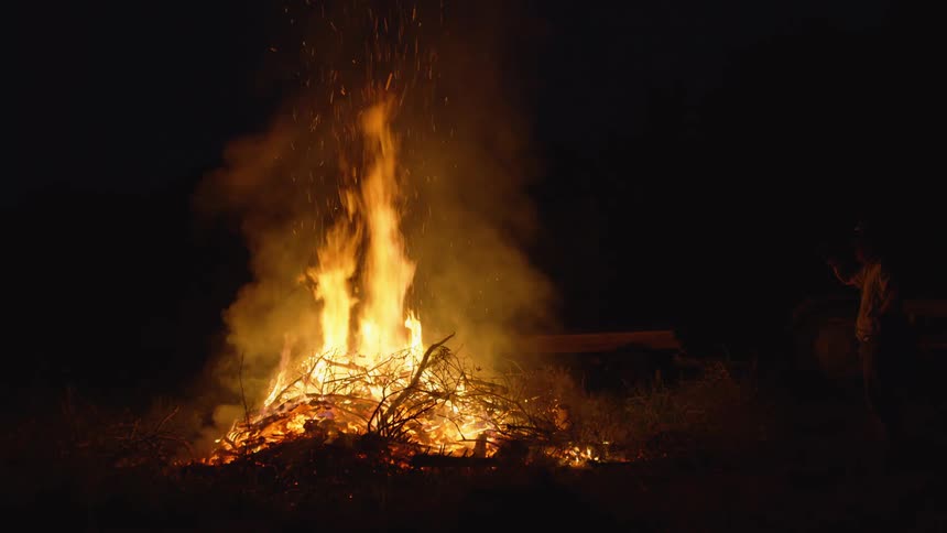 A man stands and watches a bonfire burning at night
