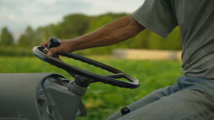 A man sits on a tractor in a rural field and starts driving it
