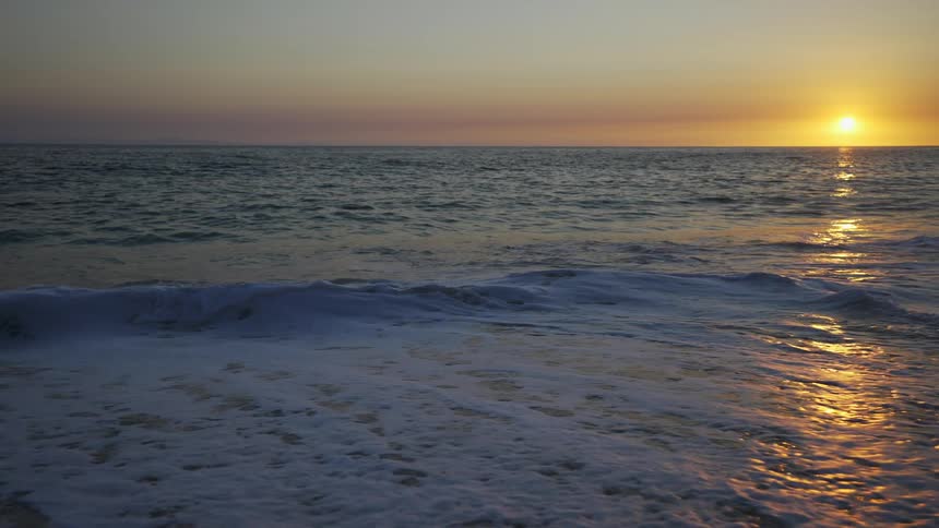 Sun nears horizon in red tinted sky reflecting off waves flowing toward and onto ocean