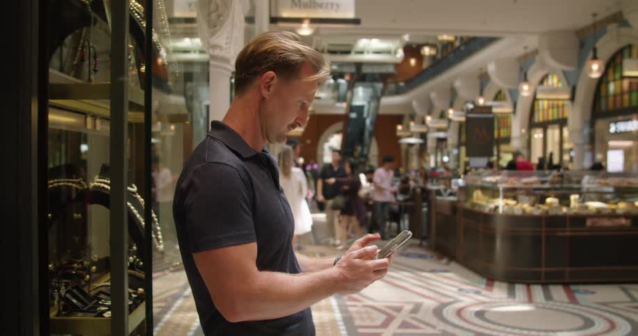 A man standing in a shopping mall types on his smartphone before walking onto an escalator