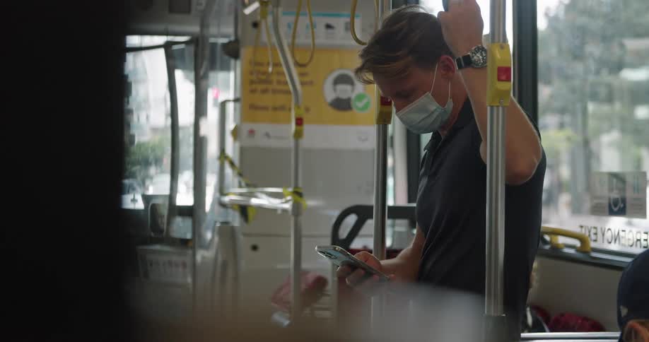Man with a dark polo shirt and light blue mask holds bus hand handle on the bus