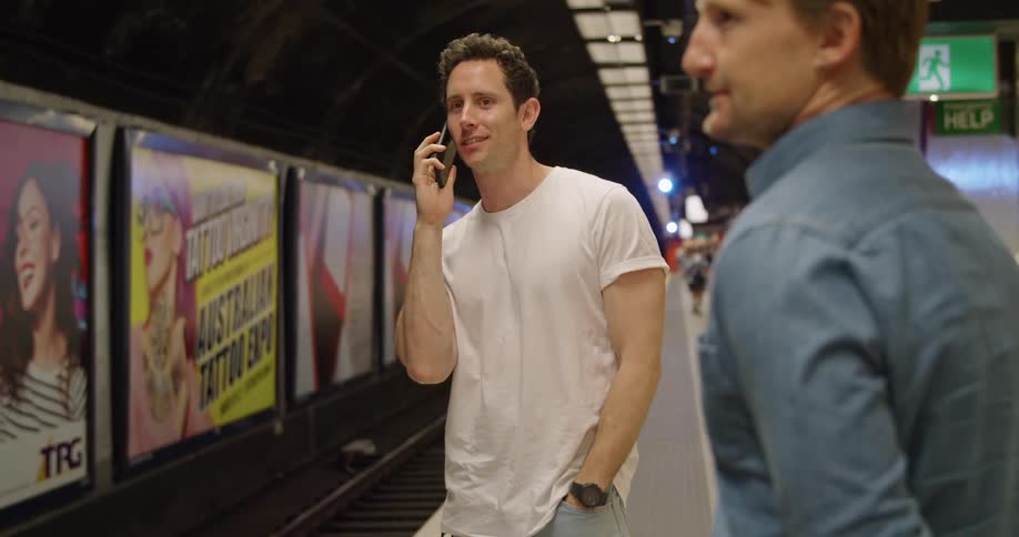 Two men in a train station with one man talking on a smartphone