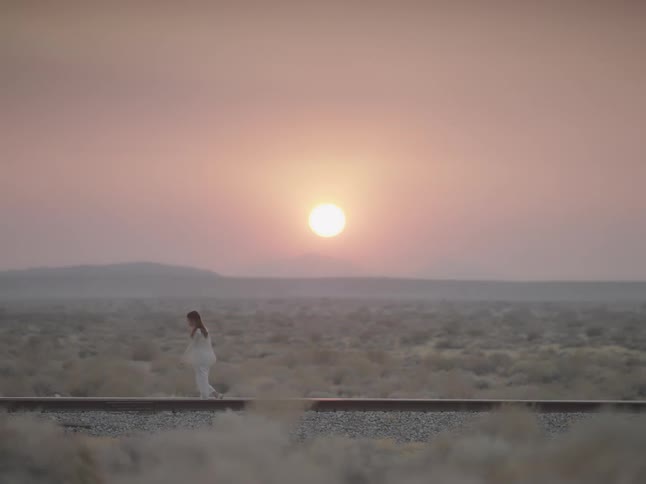 A woman walking on train tracks on a desert with the sunset the sky and clouds in the background