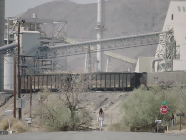 A woman walks down the middle of a road in a small town toward a refinery plant