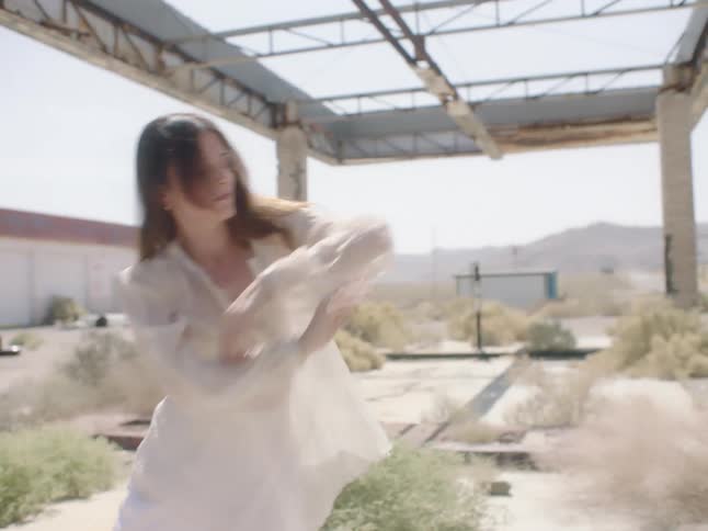 A woman wearing white pants and a see through blouse dances in a desert near buildings and hills