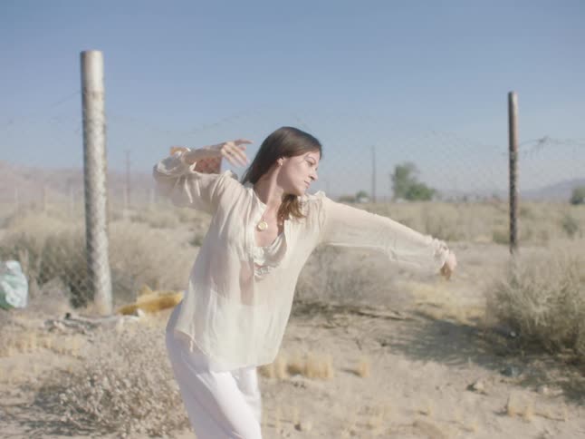 Woman wearing filmy white dances in desert near chain link fence under clear blue sky
