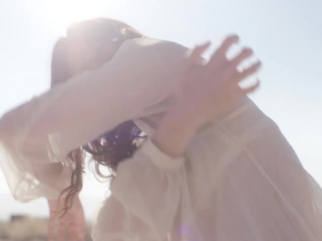 A woman kneeling in a desert dirt road dances and hugs herself