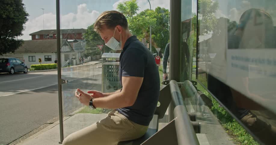 A man sits on a bench at a bus stop next to the road while checking his phone