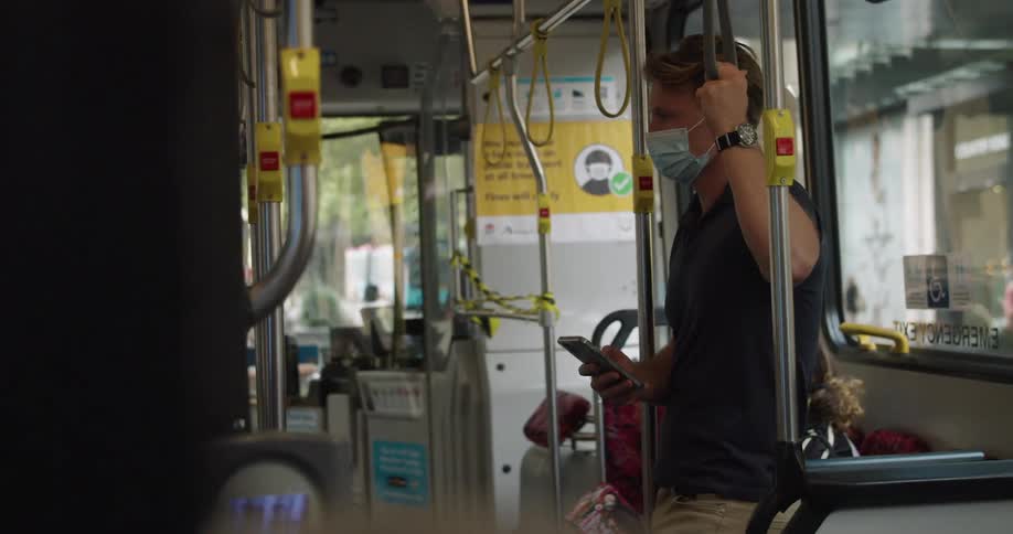 Man wearing face mask holding handle on public transportation looks around briefly before looking down at mobile phone