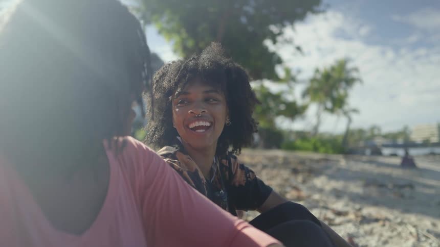 Two women sit on sand on beach laughing and talking with buildings in background