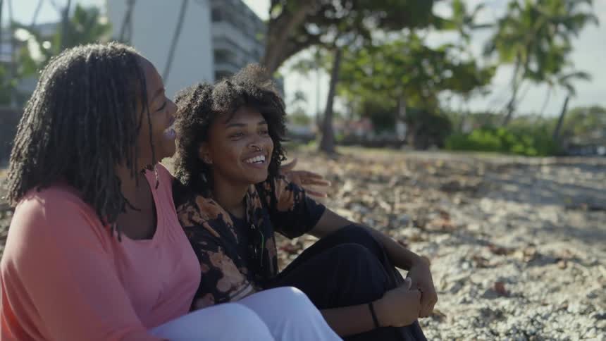Two friends smiling and laughing while sittign toghether over the sand of a beach