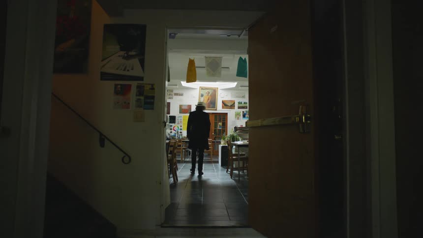 A man wearing a hat and long jacket walks through an open doorway into an art classroom