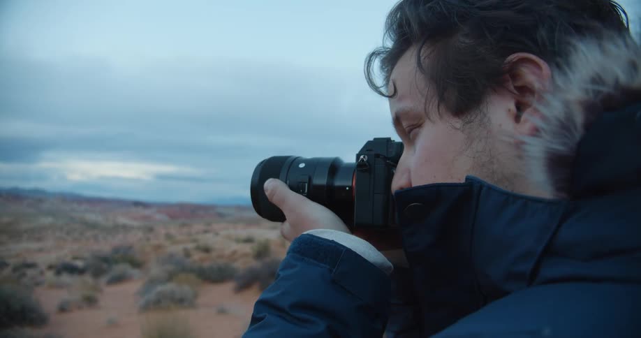 A man with a coat and a camera taking a picture with the sky and clouds in the background