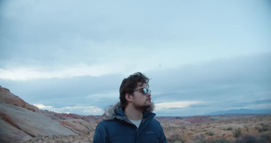 A man in a thick parka looks up at the sky as he stands and walks through the desert