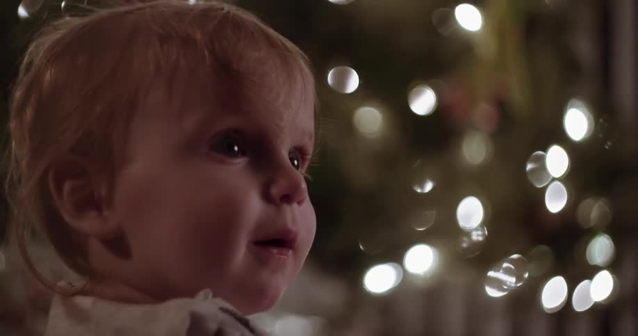 A baby looks at the lights on a christmas tree and holds up an ornament
