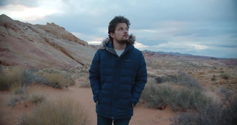 A man with a coat walking on a landscape with the sky and clouds in the background