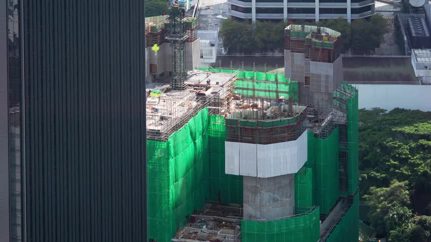 Workers move around construction site as building bordered by trees goes up next to skyscraper downtown
