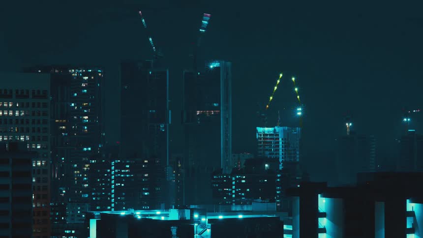Cranes on top of skyscrapers glow and shift in a city skyline at night
