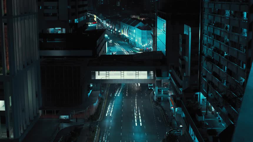 Traffic moves along city streets at night under covered pedestrian bridge connecting two buildings
