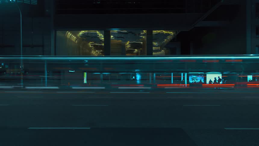 People stand and wait at a bus terminal as traffic passes by with glowing dashes of light