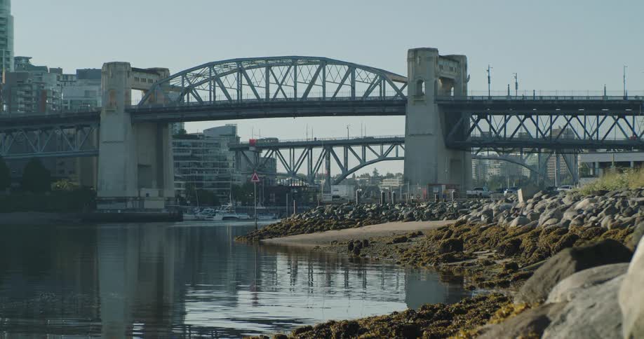 Calm water laps against sand and rocks under a Vancouver city bridge