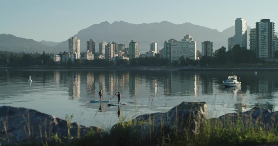 Two people row paddle boards near a city skyline