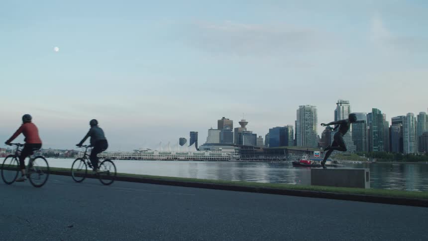 People ride their bicycles past statue in Stanley Park with the Vancouver city skyline and rising moon in the background