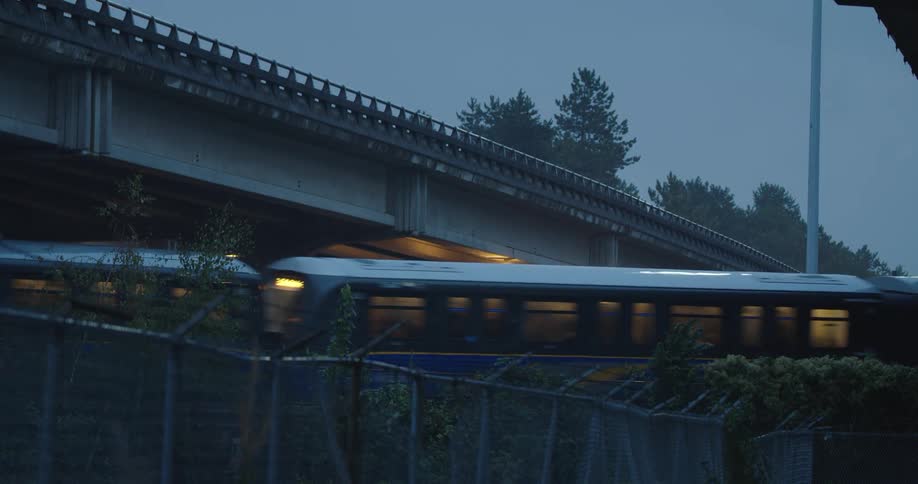 A skytrain passing under a bridge in the rain with barbed wire in front