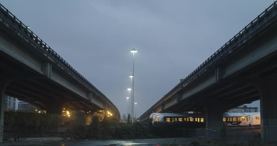 Skytrains passing underneath overpasses with streetlights on a rainy early morning