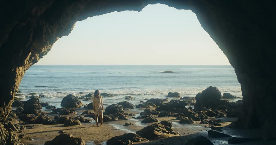 A woman standing by a cave by a rocky shore with the ocean sky and clouds in the background