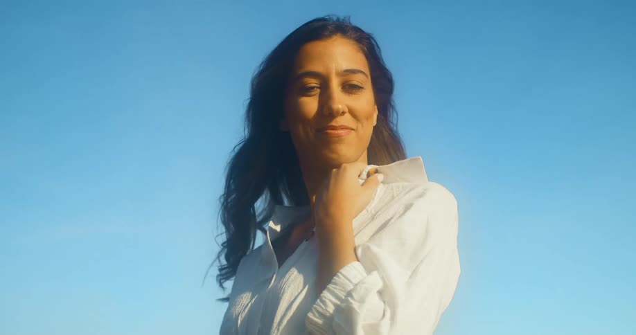 A woman in a white dress stands at the coast an smiles while posing