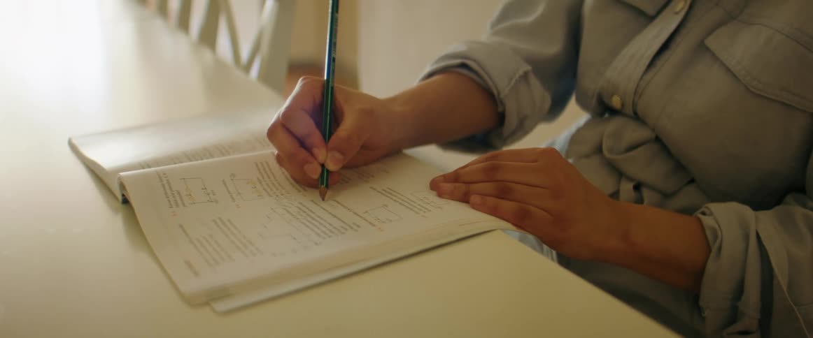 A woman sits at a table completing questions in a workbook with a pencil