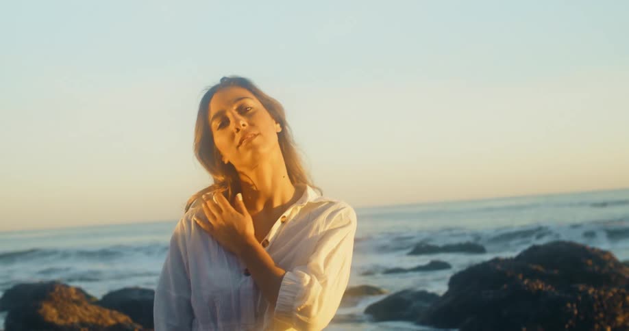 A woman poses near the ocean on a rocky shore while touching her shoulder and body