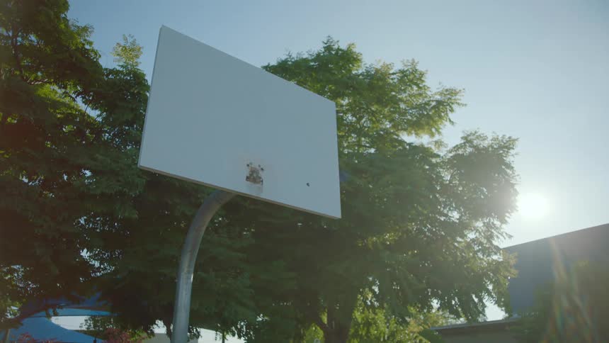 A basketball backboard in a playground with trees the sky and clouds in the background