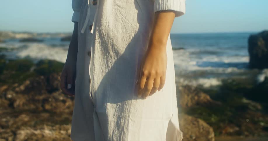 A woman stands on a rocky coastline as her clothing flutters in the wind