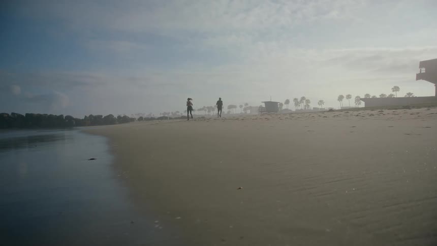 Two joggers run along beach sand near water as mist covers trees on horizon