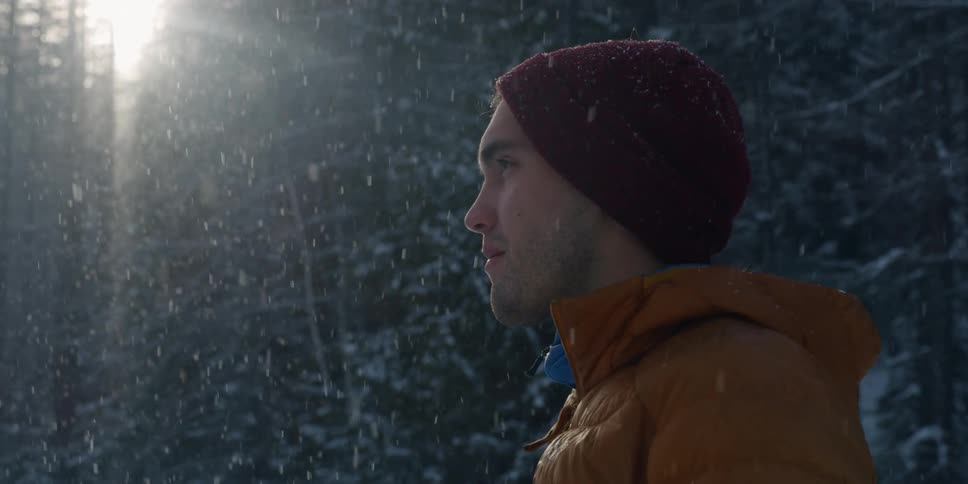A man wearing a beanie and a heavy coats stands outside as the snow falls