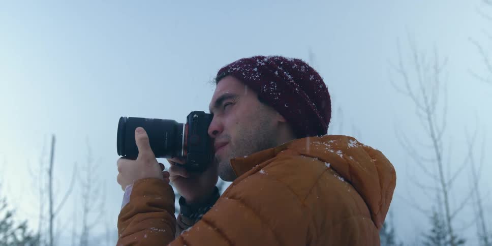 A man squints as he looks through a camera lens taking a photo in the snow