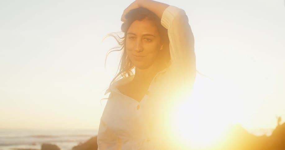 The sun glares behind a woman smiling and posing near the ocean